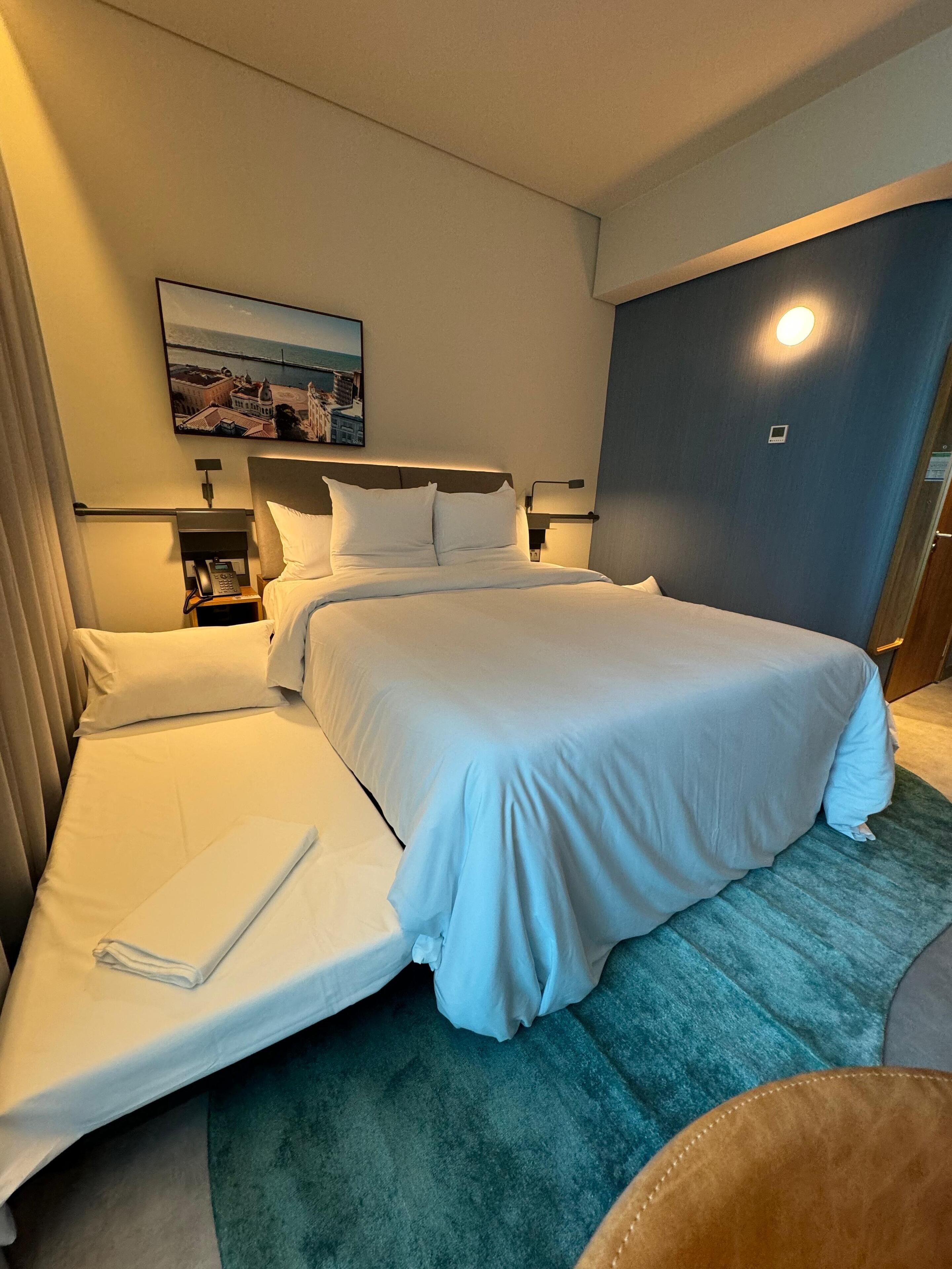 Quarto de hotel com uma cama grande, mesa de cabeceira com abajur, telefone e um quadro na parede.