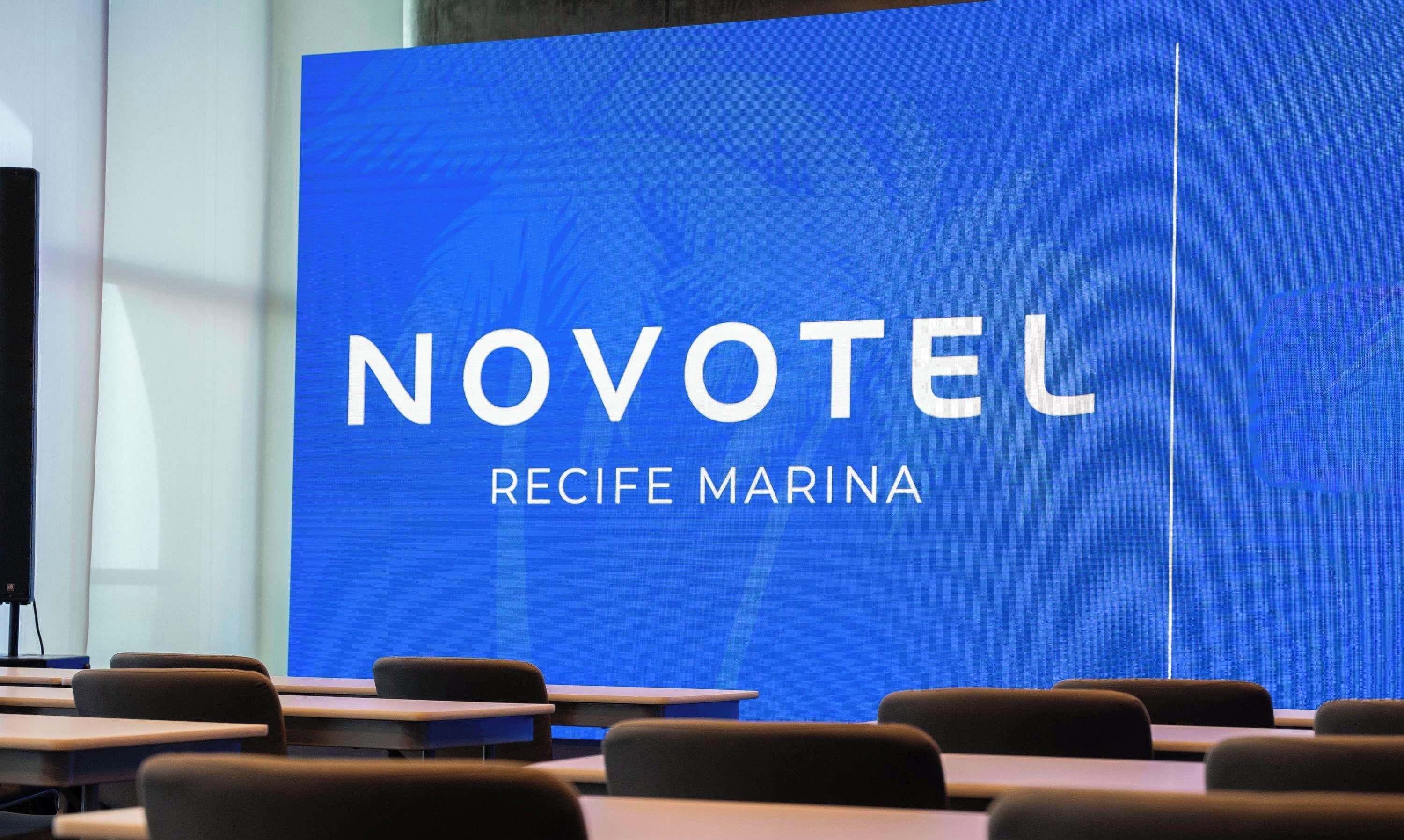 Uma sala de conferências com uma tela grande exibindo 'NOVOTEL RECIFE MARINA'.