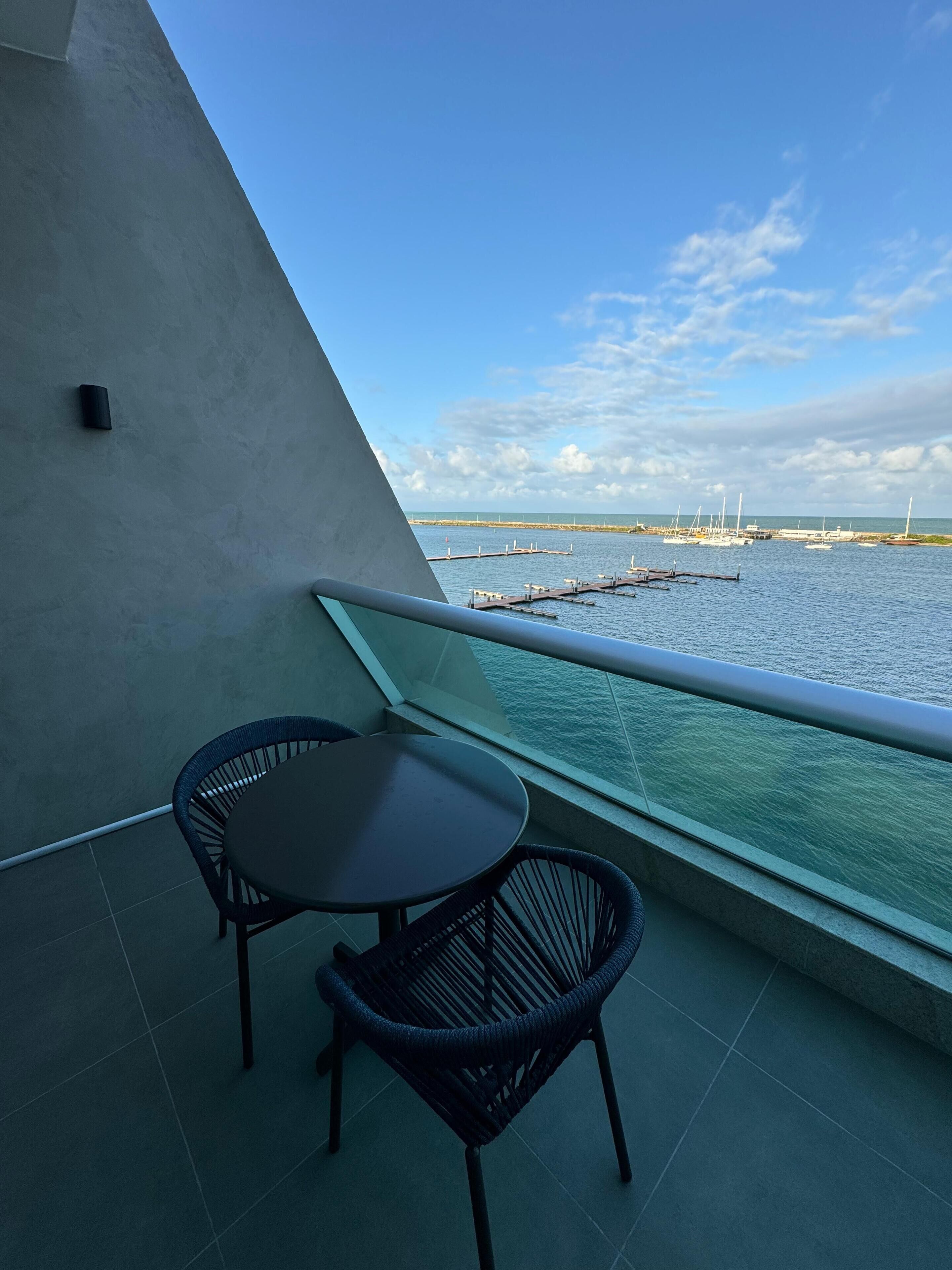 Un balcon avec une table et des chaises, donnant sur une marina où sont amarrés des bateaux, et sous un ciel dégagé.