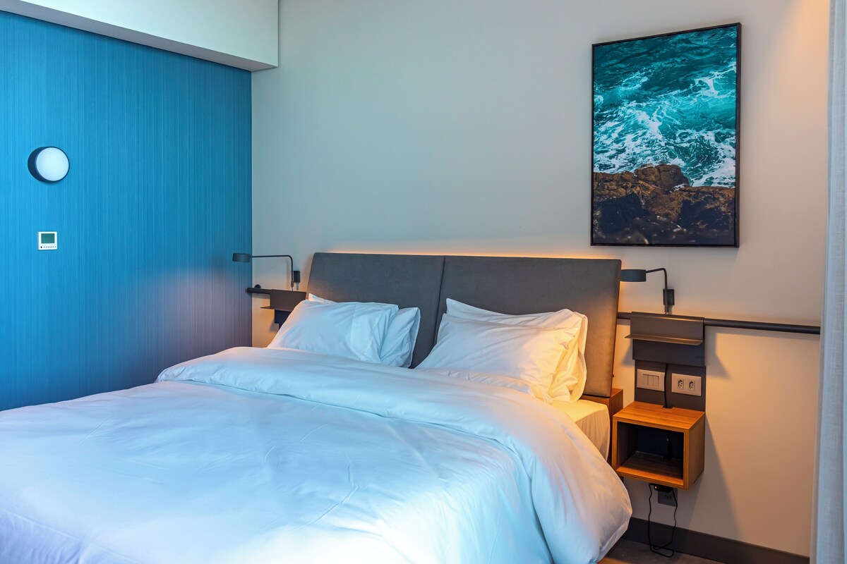 Una cama bien tendida con sábanas blancas, cabecero, mesitas de noche con lámparas y un cuadro colgado en la pared que representa olas marinas.