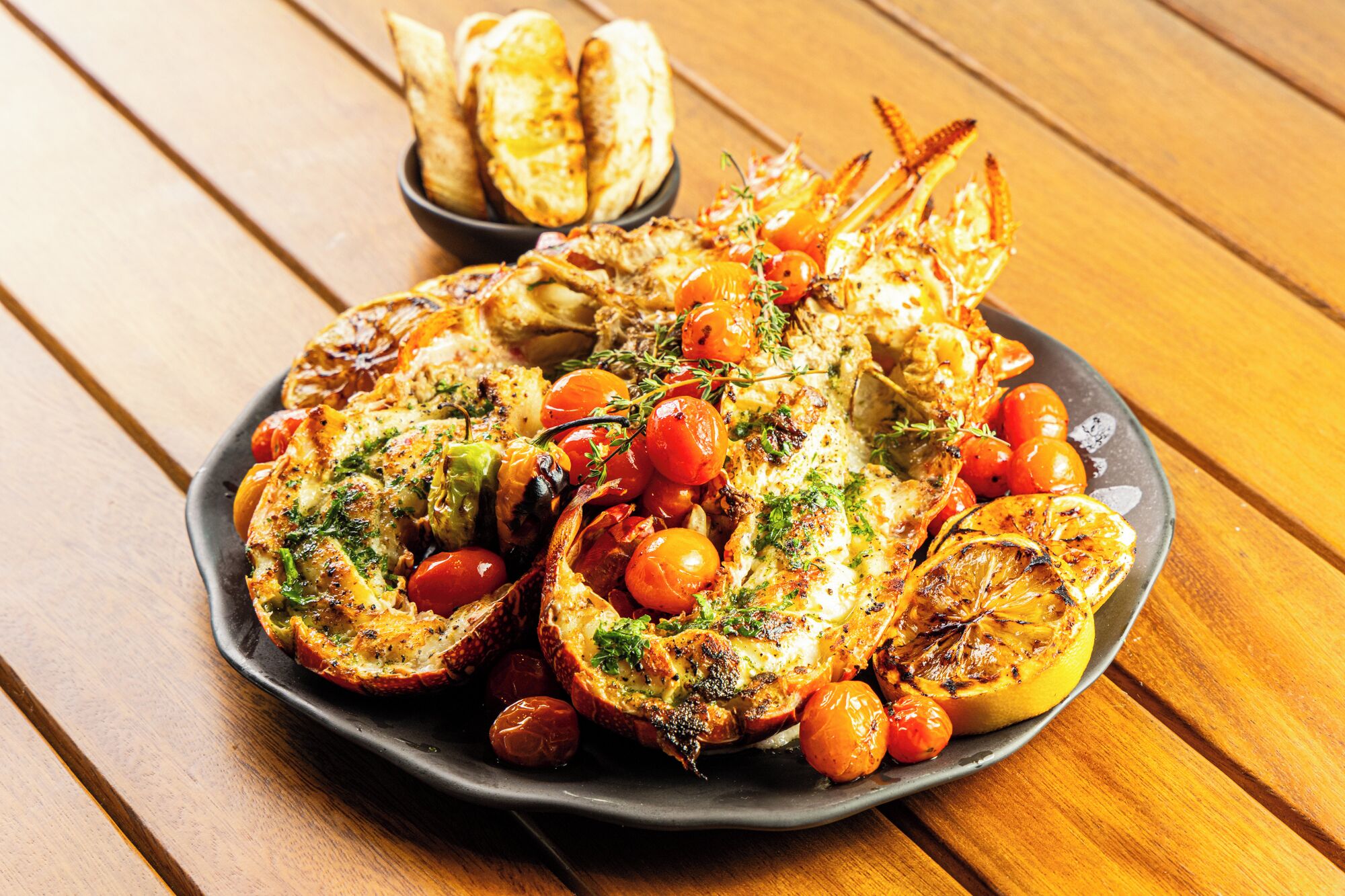 Un plato de mariscos a la parrilla con tomates y hierbas, acompañado de pan sobre una mesa de madera.