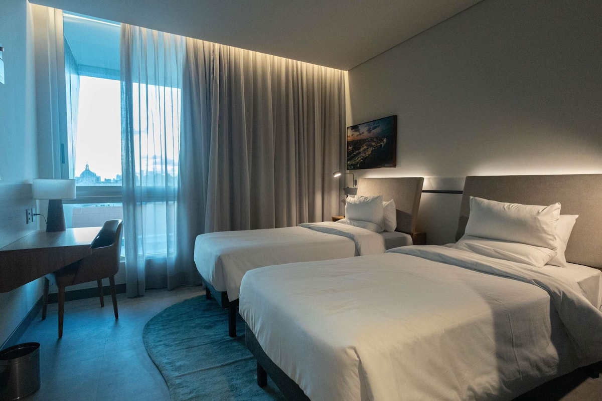 Ein Hotelzimmer mit zwei Betten, einem Schreibtisch, einem Sessel und Blick auf eine Stadtlandschaft.