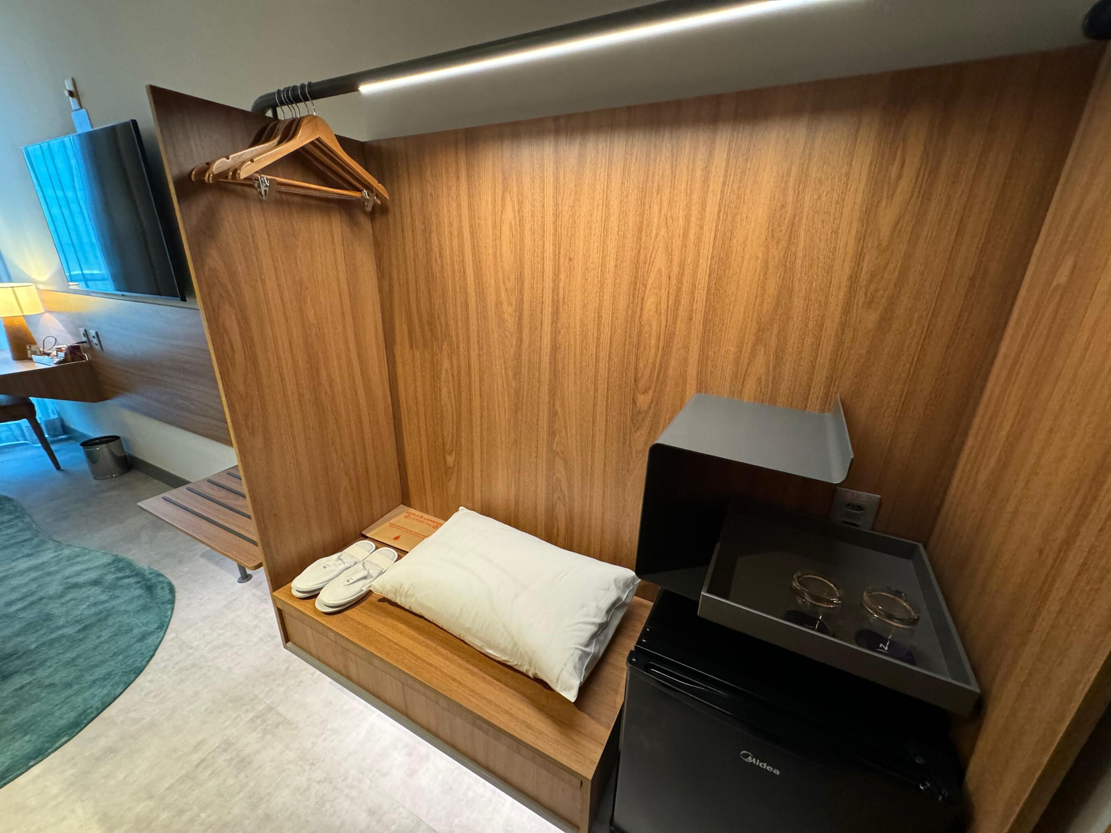 Una habitación compacta con una cama, una mesita con lámpara, un armario de madera y un electrodoméstico negro.
