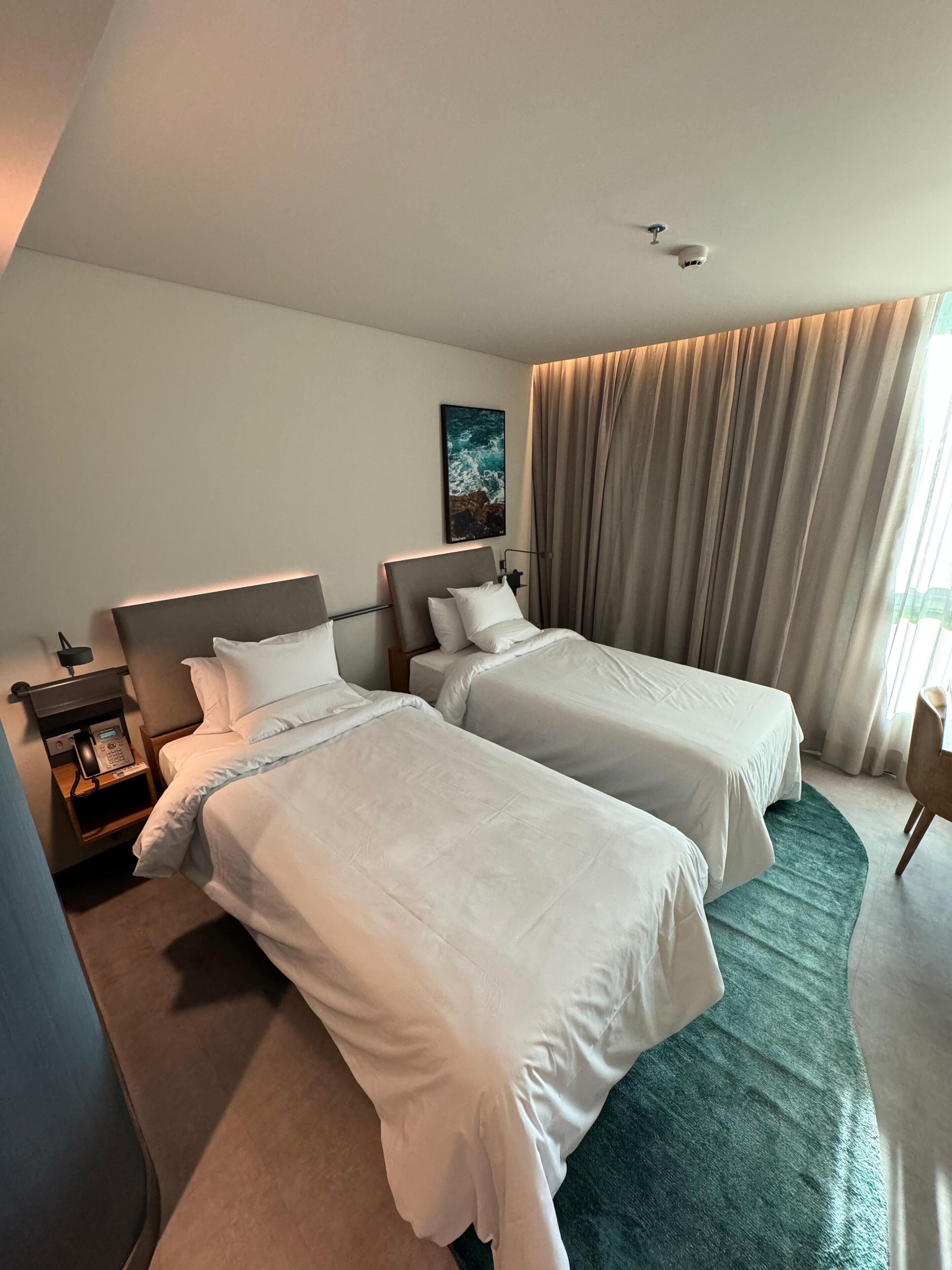 Una habitación de hotel con dos camas, una mesita de noche con teléfono, un cuadro en la pared y una ventana con cortinas.
