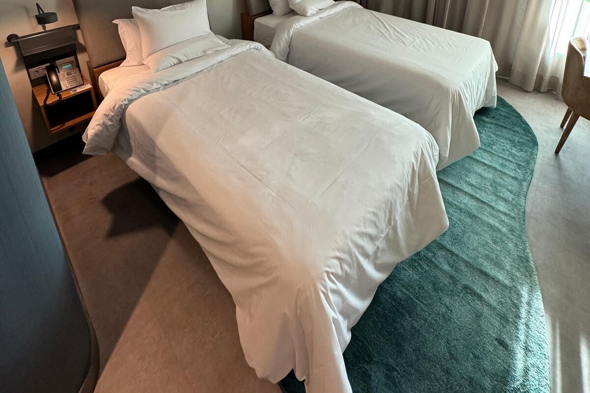 Una habitación de hotel con dos camas, una mesita de noche con teléfono, un cuadro en la pared y una ventana con cortinas.