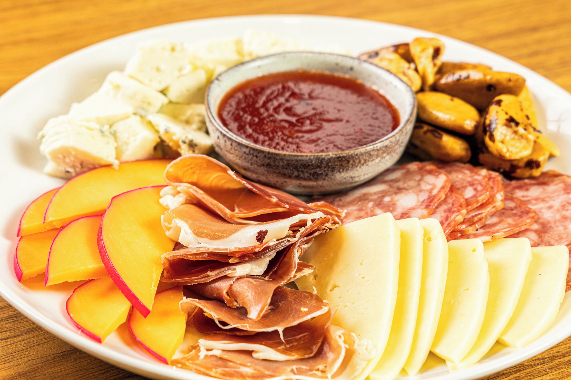 Una bandeja con quesos variados, embutidos y frutas, acompañada de un tazón de mermelada.