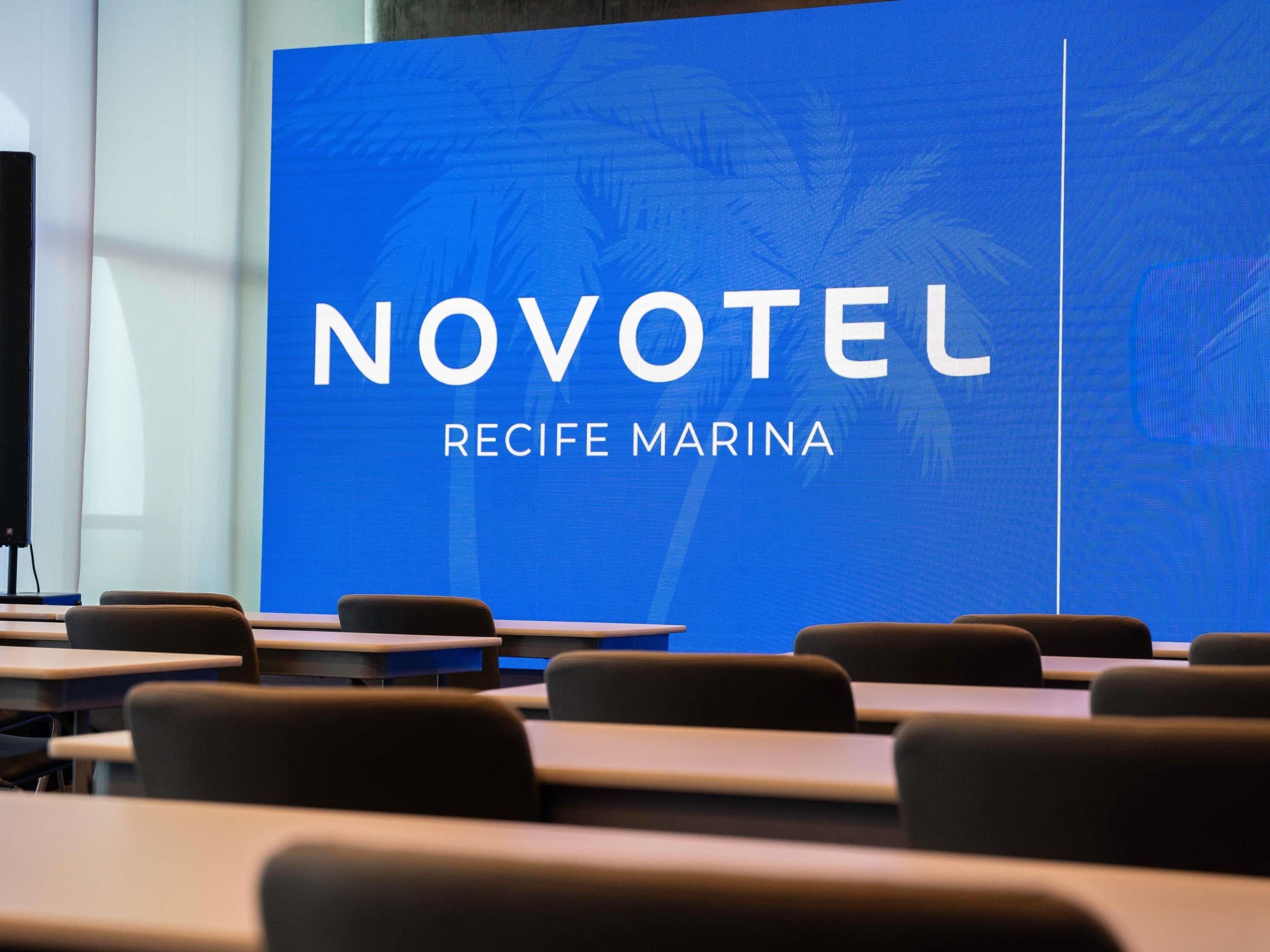 Un salón de conferencias con una pantalla grande que muestra 'NOVOTEL RECIFE MARINA'.