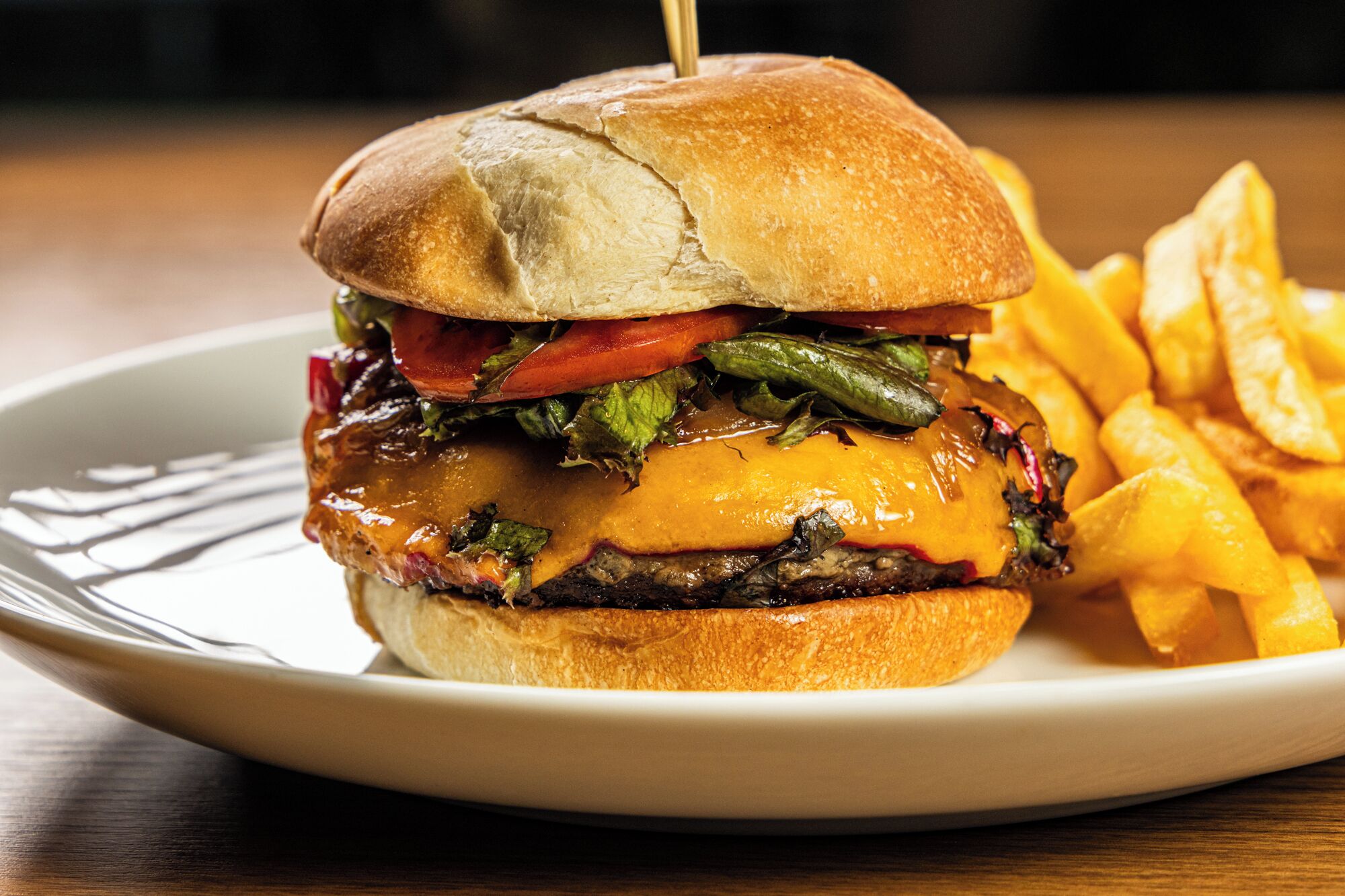 A burger with a bun, lettuce, tomato, and cheese, served with fries on a white plate.