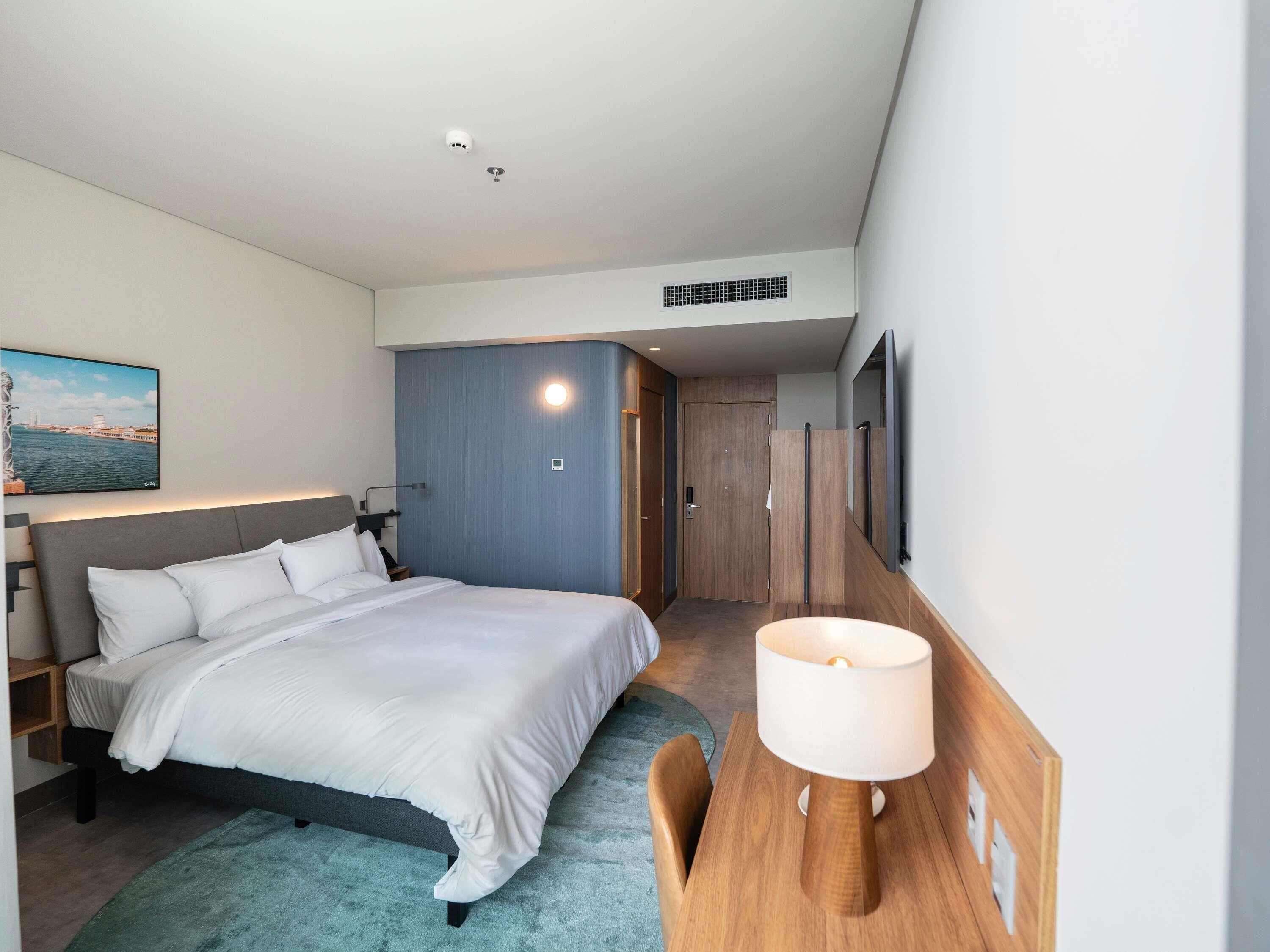 A modern hotel room with a bed, bedside table, lamp, and a TV mounted on the wall.
