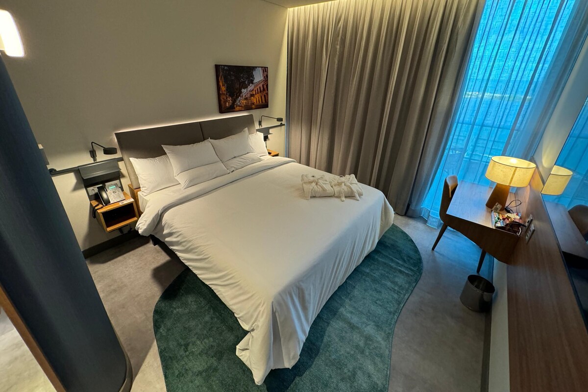Habitación de hotel con una cama grande, un escritorio con lámpara, una mesita de noche con teléfono y un cuadro en la pared.