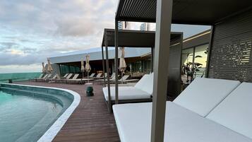 Uma área na beira da piscina com espreguiçadeiras e guarda-sóis, um deck de madeira e vista para o oceano.