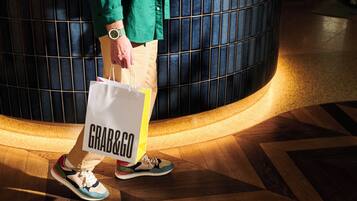Una persona sosteniendo una bolsa 'Grab&Go', de pie sobre un suelo estampado con un elemento arquitectónico circular al fondo.