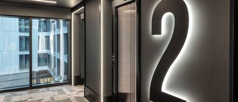 A modern elevator with a large illuminated number "2" on the wall, a patterned carpet, and a view of a building through glass doors.
