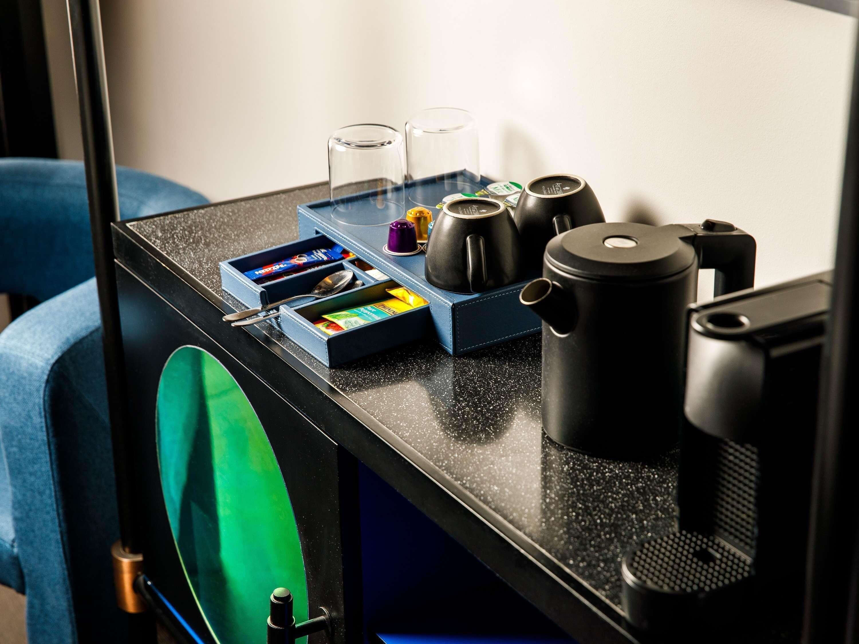 Una encimera de cocina con una cafetera, un hervidor, una tetera, una bandeja azul con utensilios y una caja de bolsitas de té.