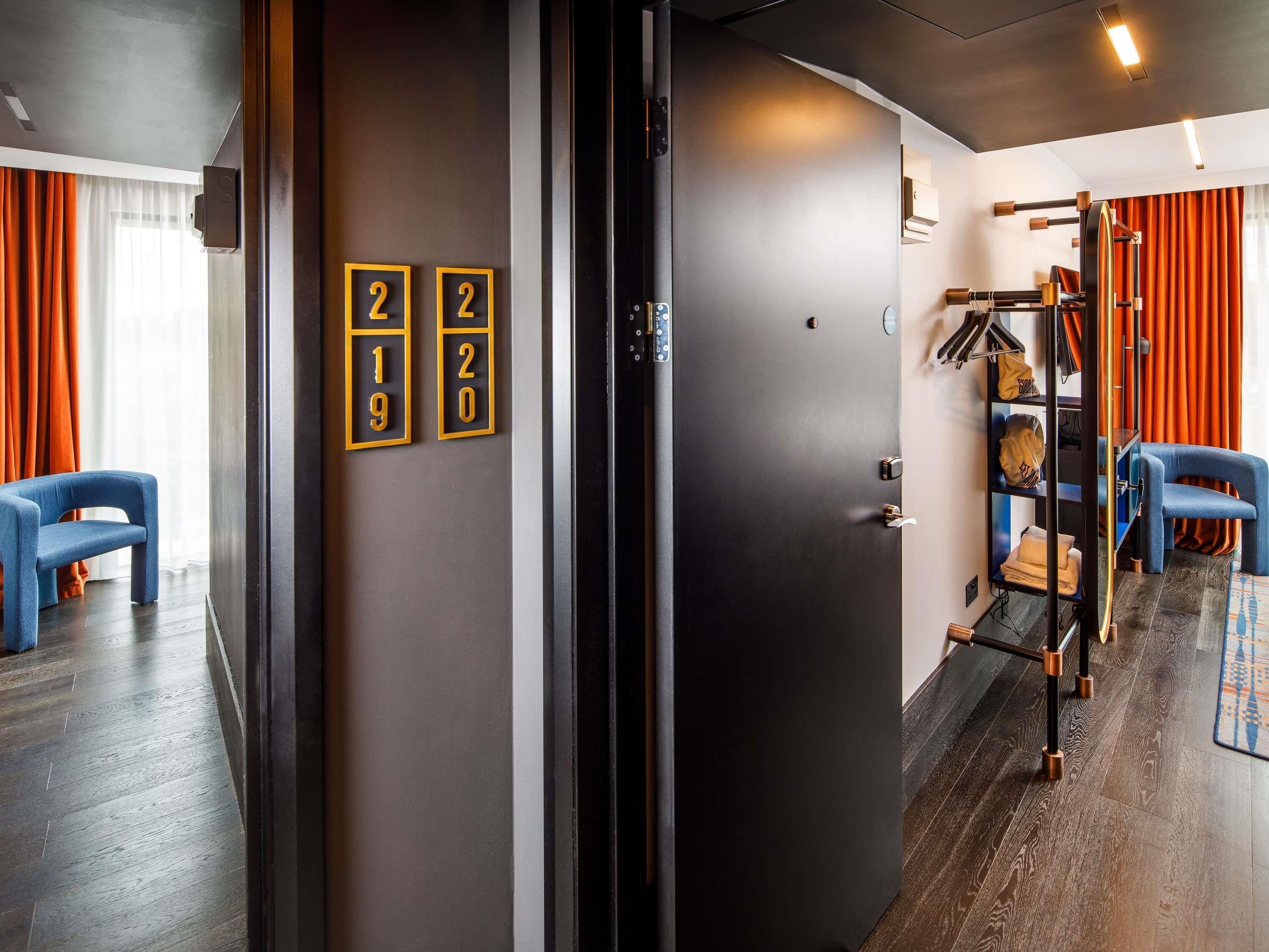 Ein Hotelzimmer mit dunklem Holzboden, einem blauen Sessel, einem Kleiderschrank mit Kleiderstange und einer Tür mit Raumnummern.