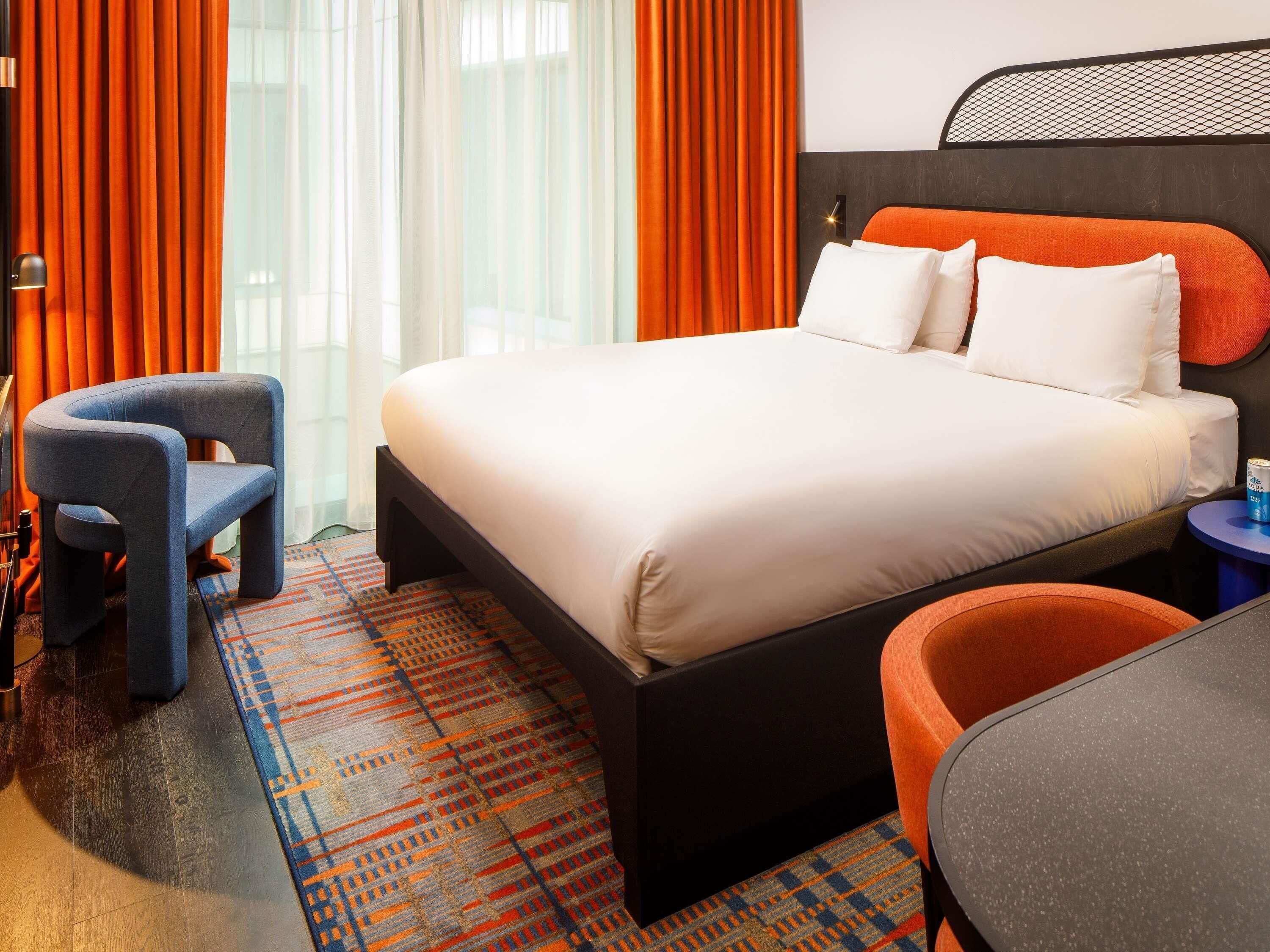A hotel room with a bed, two chairs, a small table, a patterned rug, and orange curtains.