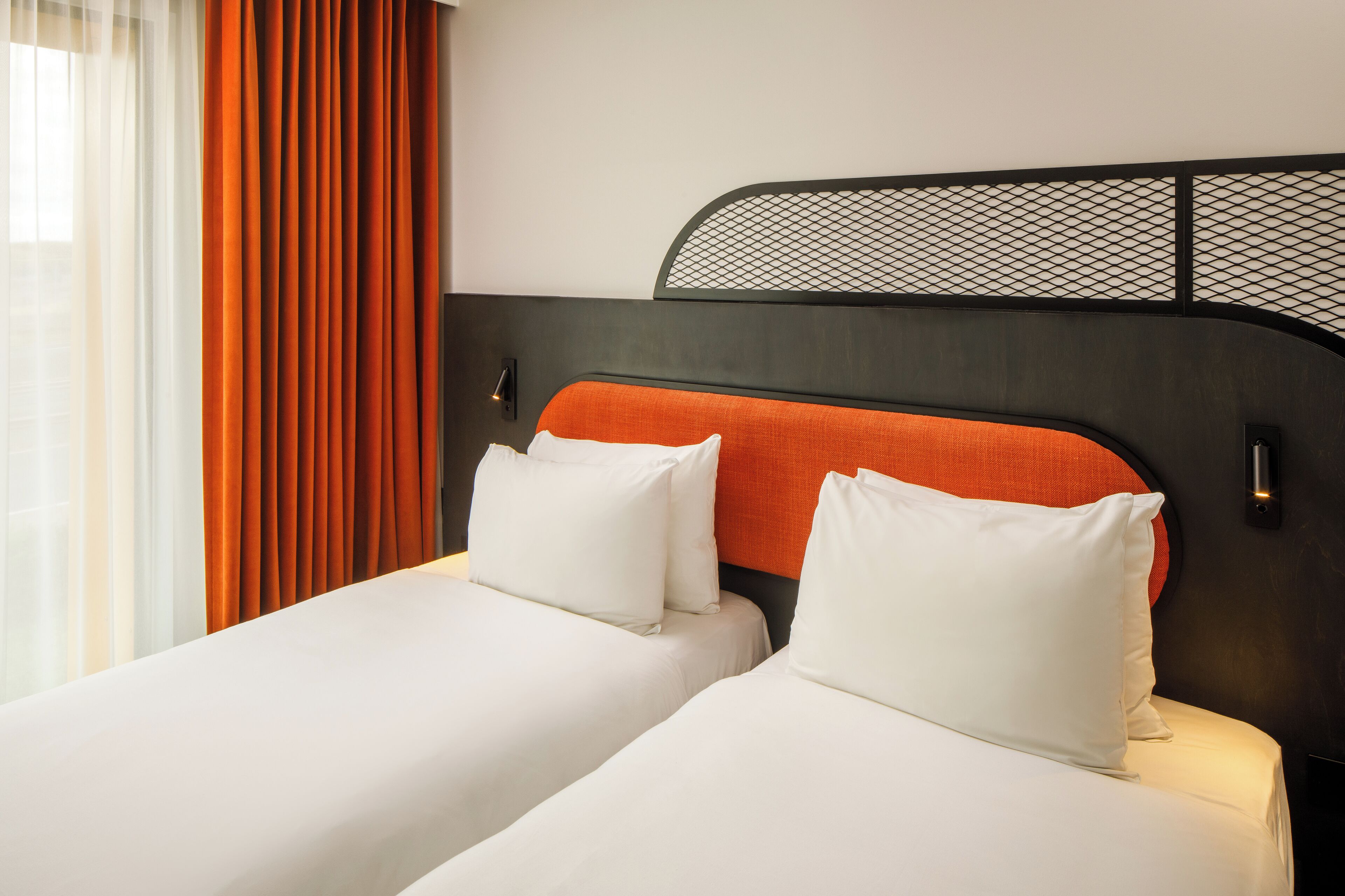 Cama doble con ropa blanca y cabecera naranja, dos almohadas blancas y cabecera de malla negra.