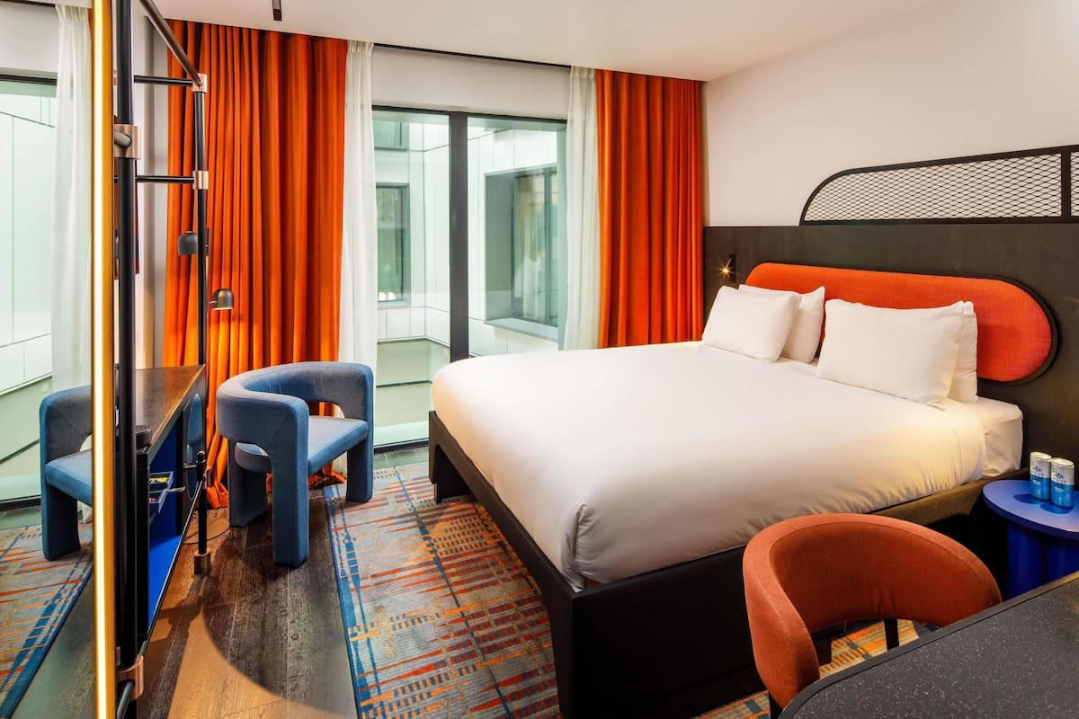 Una habitación de hotel moderna con una cama grande, dos sillones, una mesita y vistas a la ciudad a través de amplios ventanales.