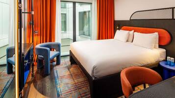 A modern hotel room with a large bed, two armchairs, a small table, and a view of the city through large windows.