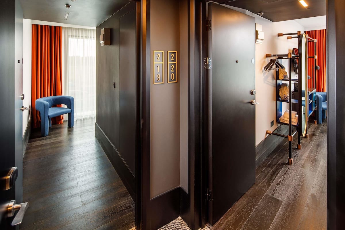 Habitación de hotel con piso de madera, una silla azul, un armario y un perchero con objetos colgados.