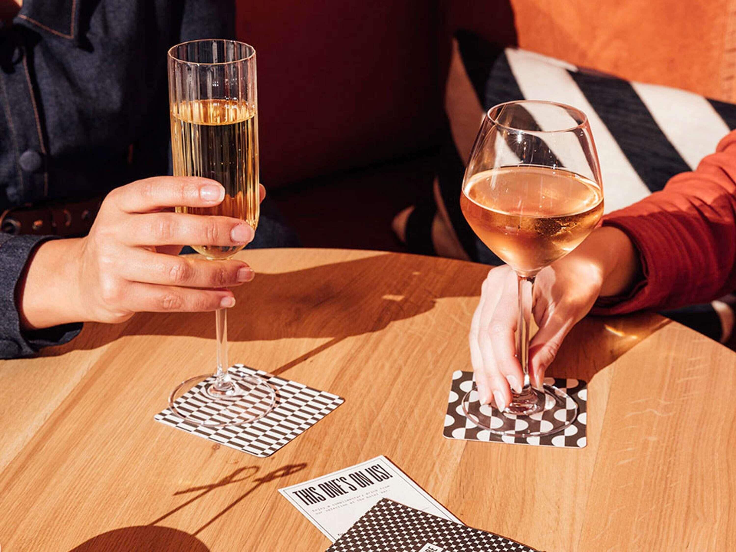 Twee personen met wijn glazen aan een houten tafel, met geruit tafelonderzetters en een kaartspel.