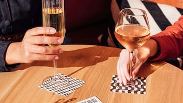 Dos personas sosteniendo copas de vino en una mesa de madera con posavasos de cuadros y un juego de cartas.