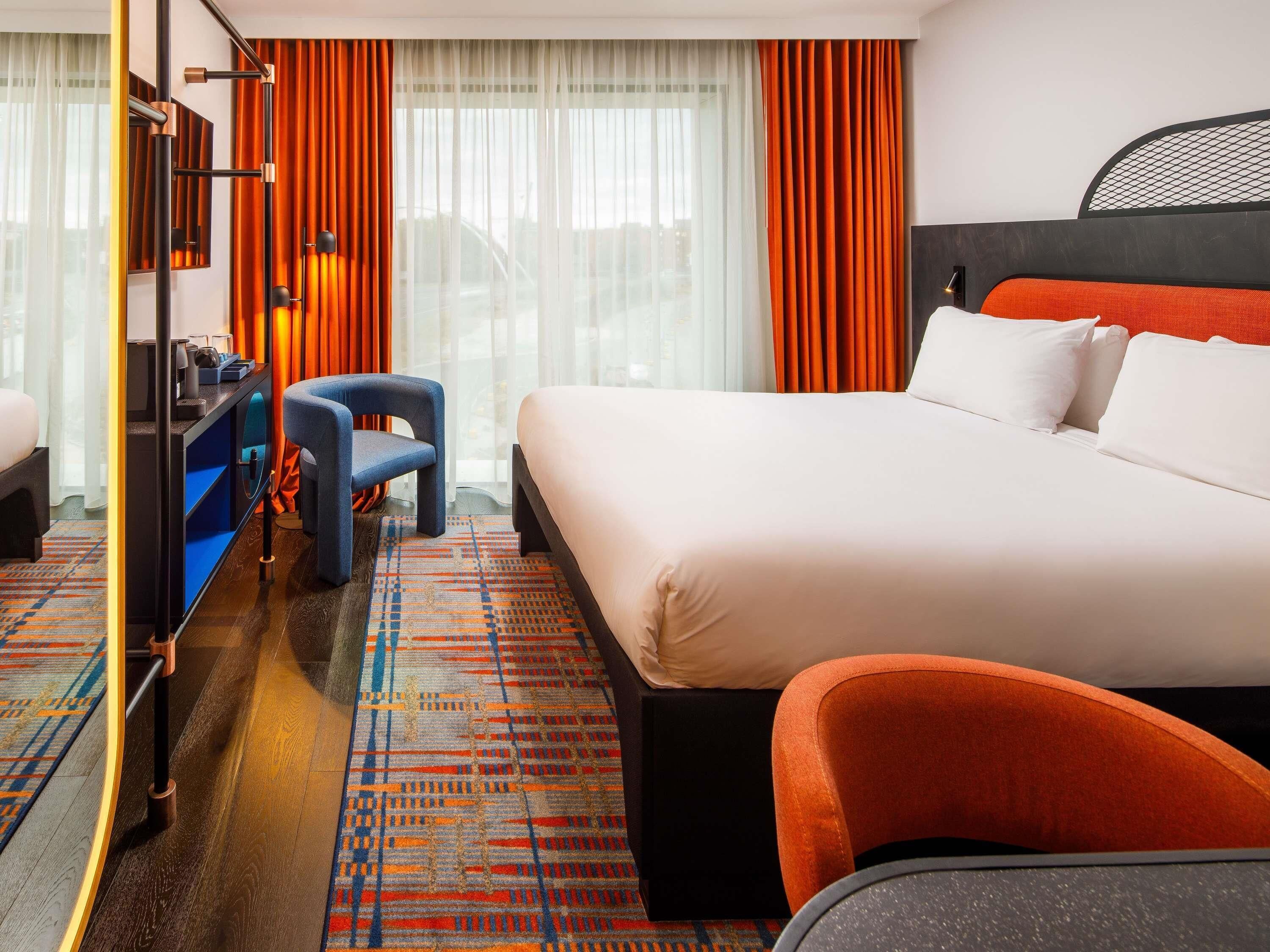 Habitación de hotel con una cama grande, un escritorio, una silla y vistas a la ciudad a través de amplias ventanas.