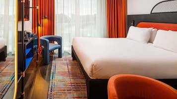 Habitación de hotel con una cama grande, un escritorio, una silla y vistas a la ciudad a través de amplias ventanas.