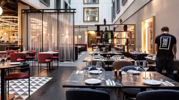 A modern restaurant with a central dining area, a bar, and a bookshelf. - TRIBE Manchester Airport (Manchester)