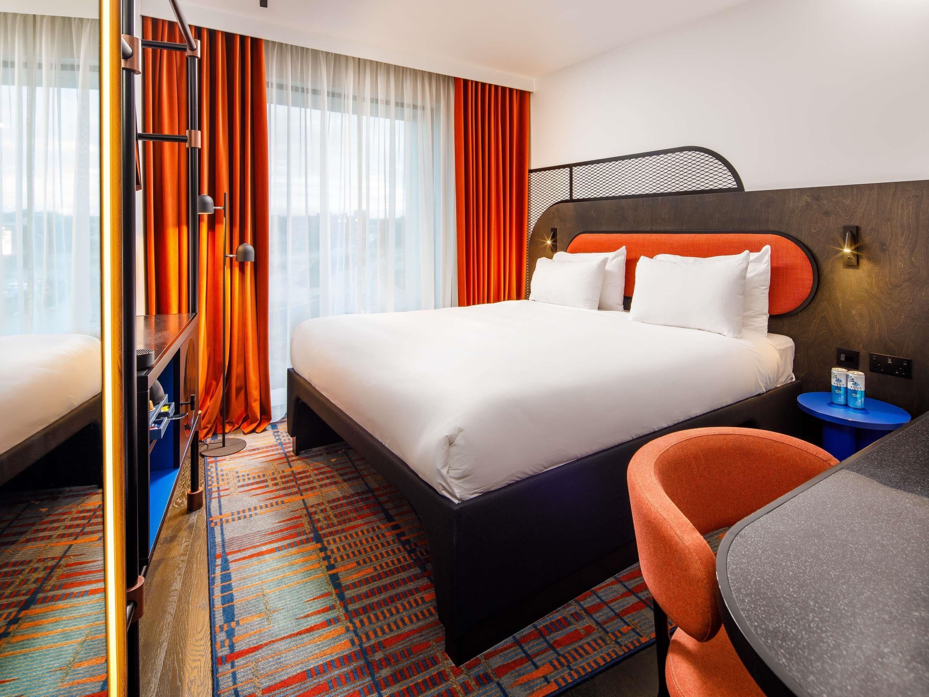 Hotelkamer met een groot bed, een bureau met twee blikjes bier, een stoel en een kamerbreed tapijt met een patroon.