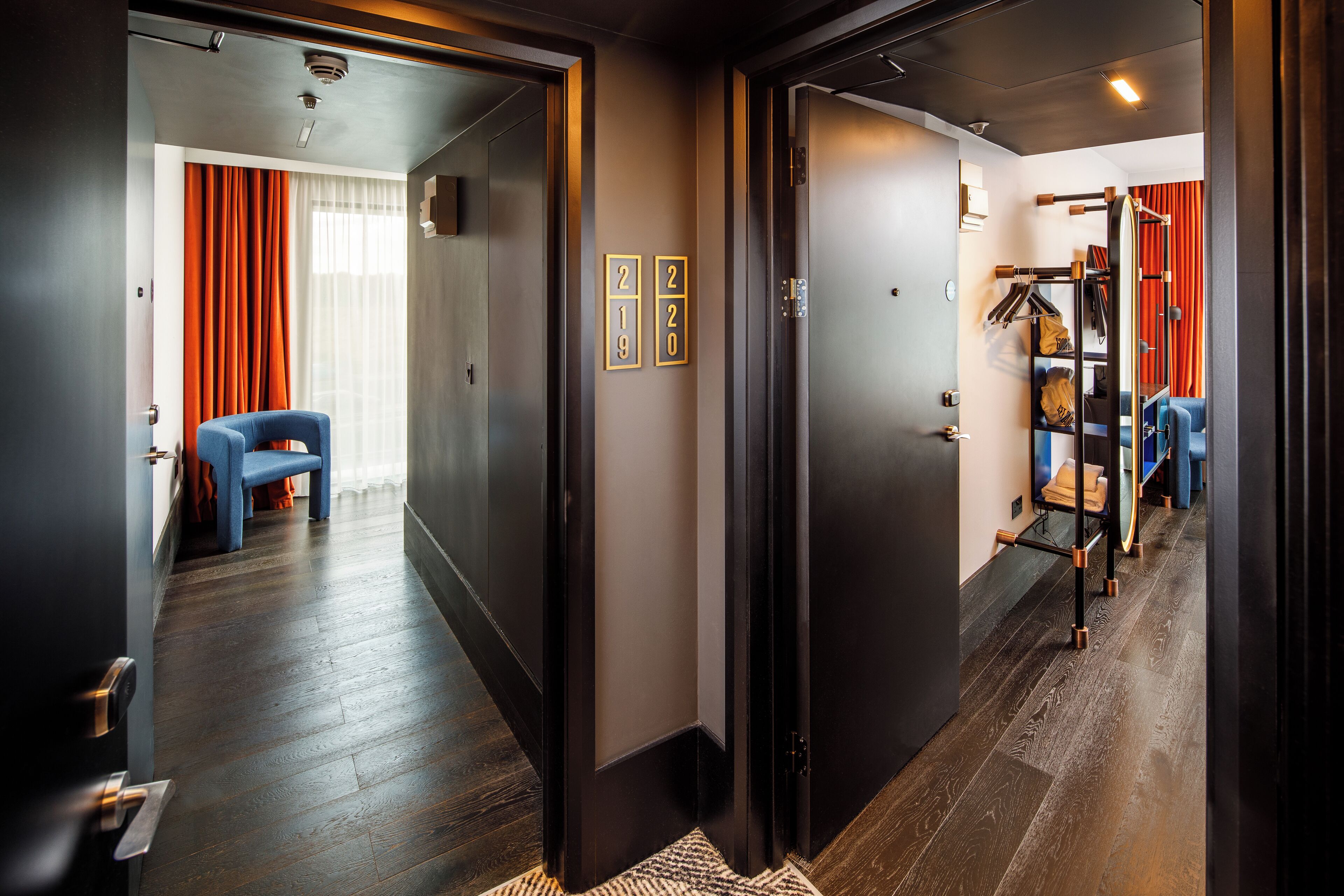 A hotel hallway with dark wooden floors, a blue chair, and a coat rack with hanging items.