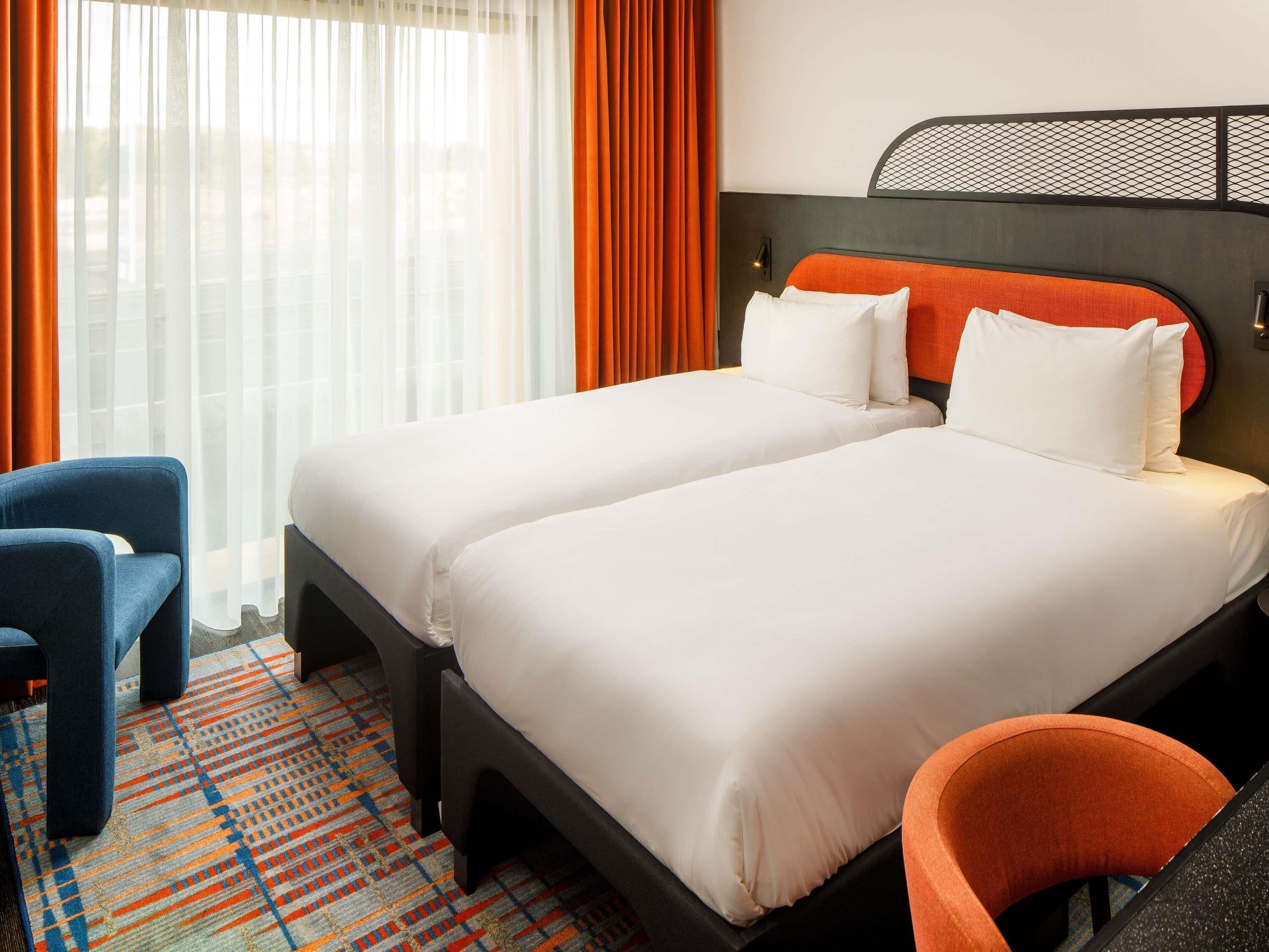 A hotel room with two beds, a blue armchair, an orange armchair, and a patterned carpet.