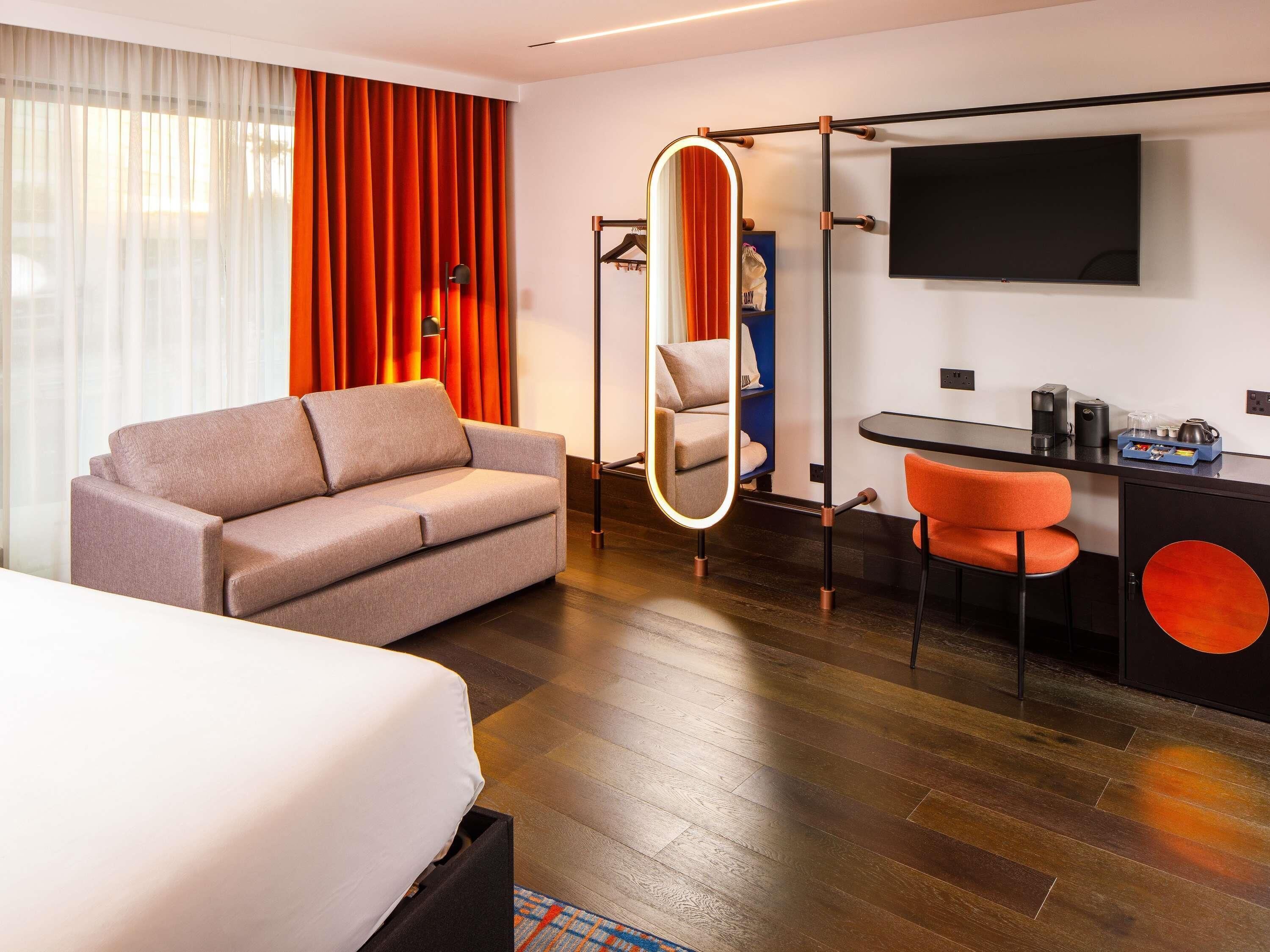 A modern hotel room with a sofa, a bed, a desk with a chair, a TV, and a mirror.
