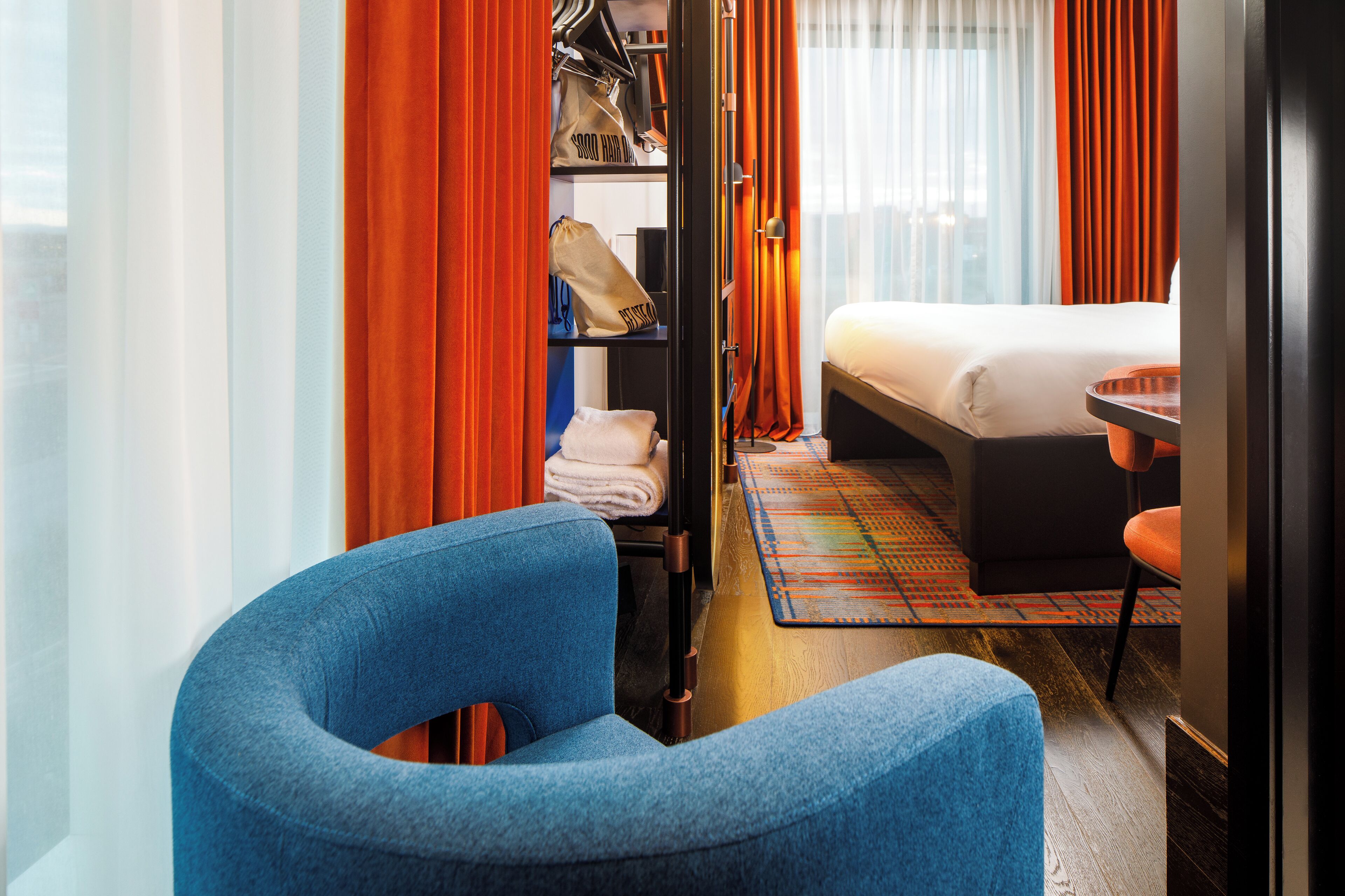 A hotel room with a bed, a blue armchair, a small table, and a wardrobe with hanging clothes and bags.