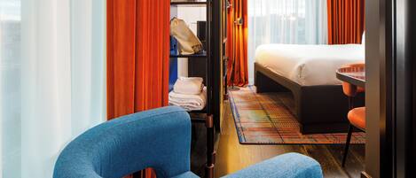 A hotel room with a bed, a blue armchair, a small table, and a wardrobe with hanging clothes and bags.