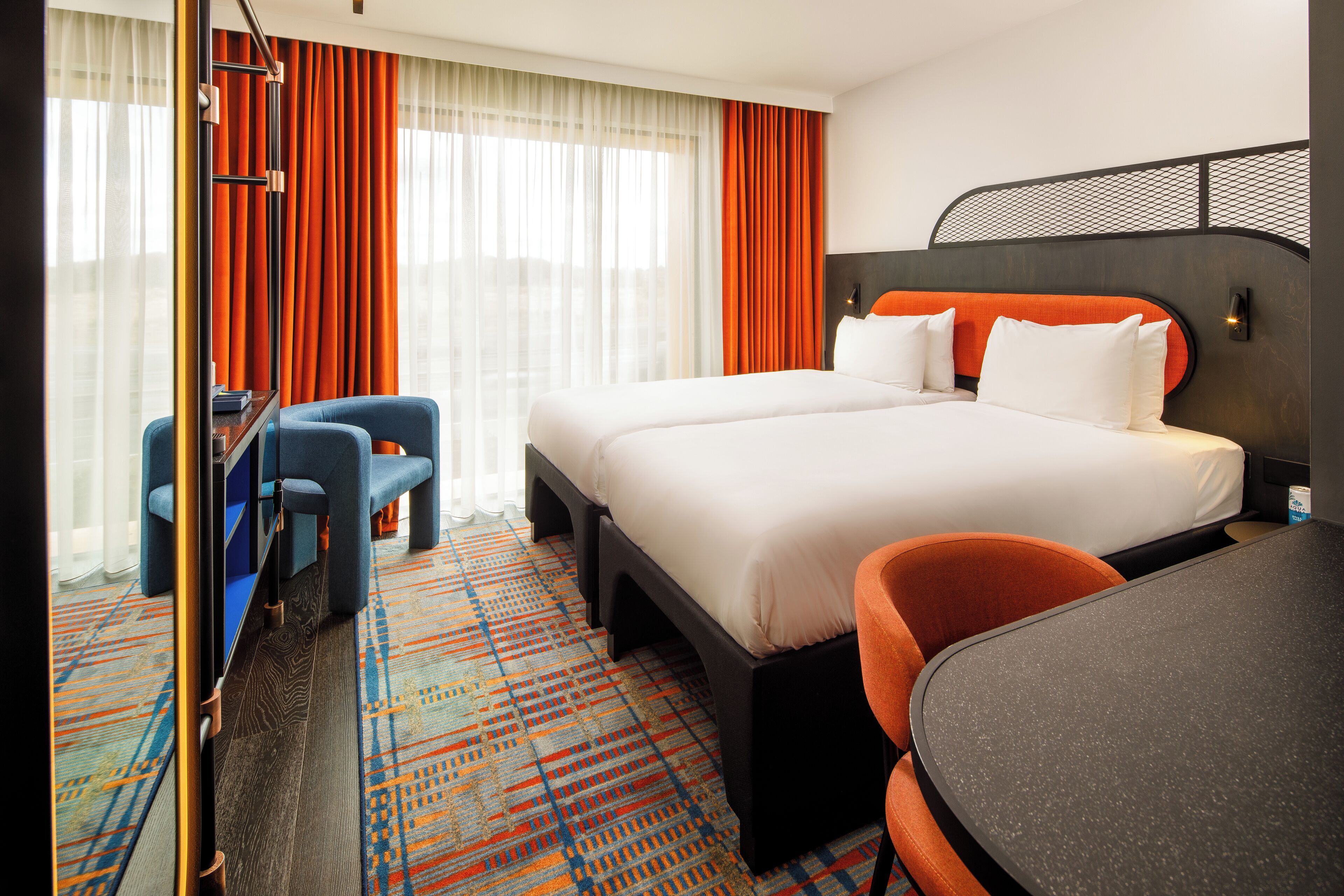 A hotel room with a large bed, two chairs, a desk, and a patterned carpet.