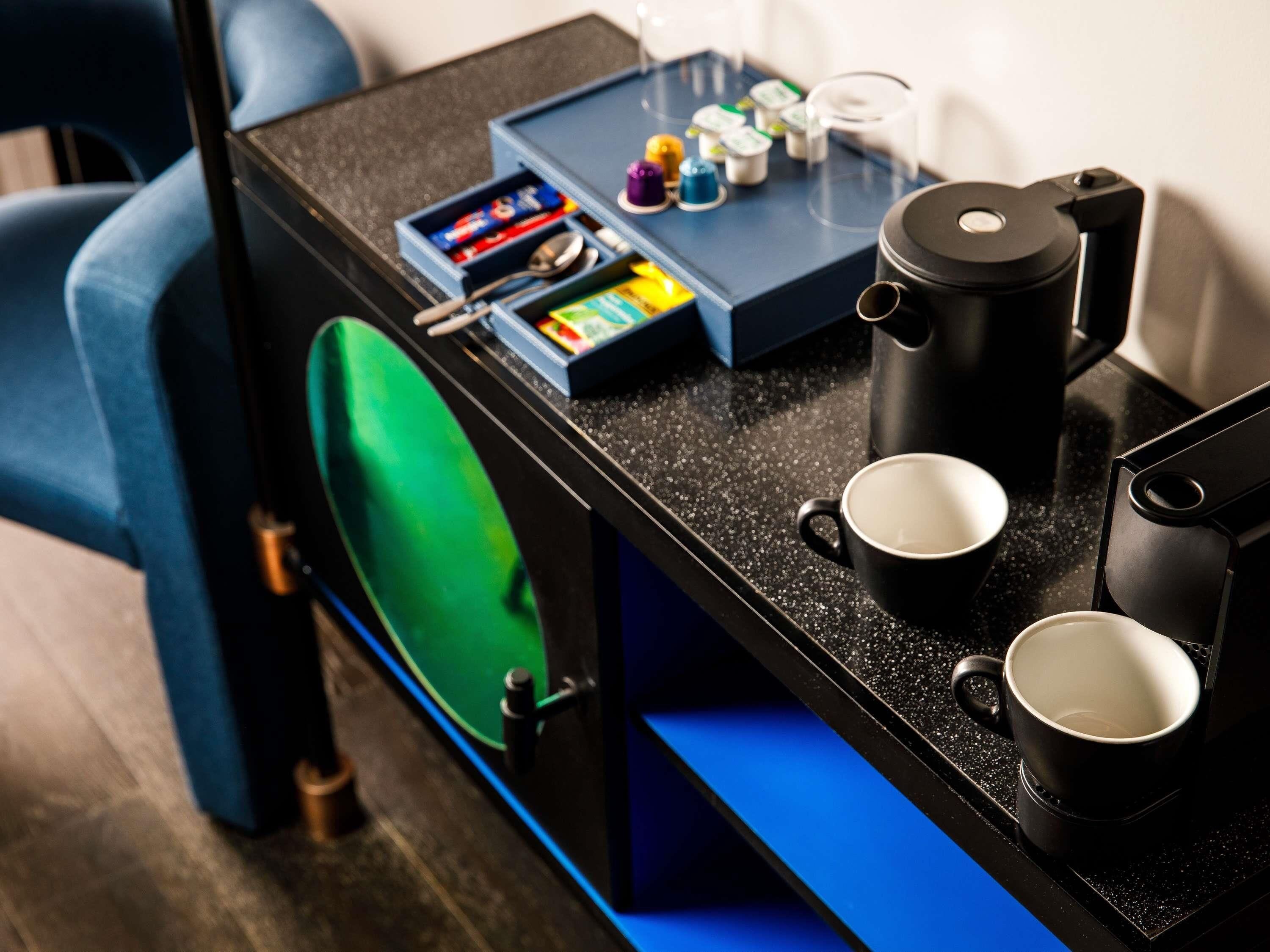 A compact kitchen area with a coffee maker, two mugs, a tea set, and a small table with various items.