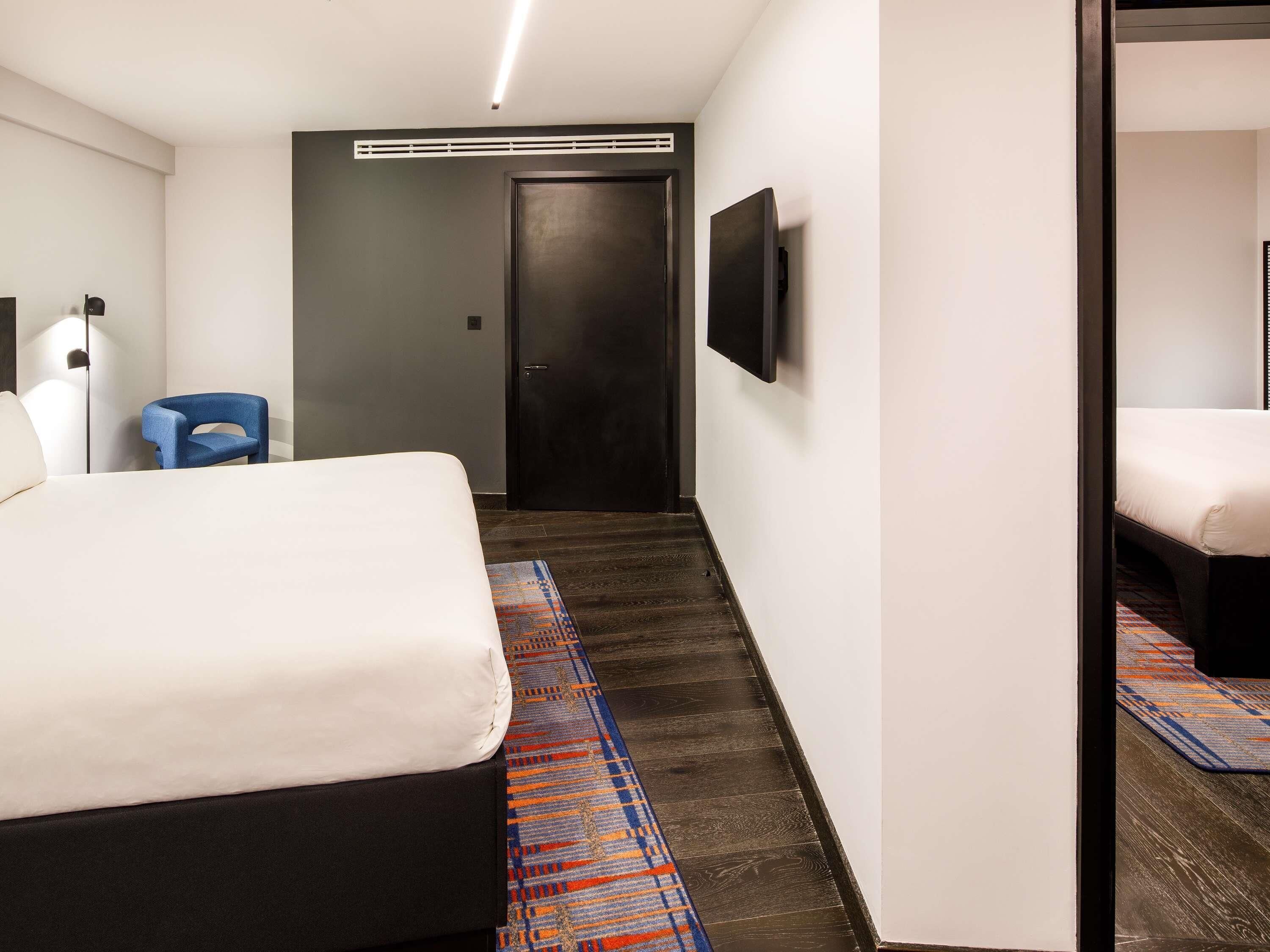 Una habitación de hotel con cama, una silla azul, un televisor y una puerta que da a otra habitación.