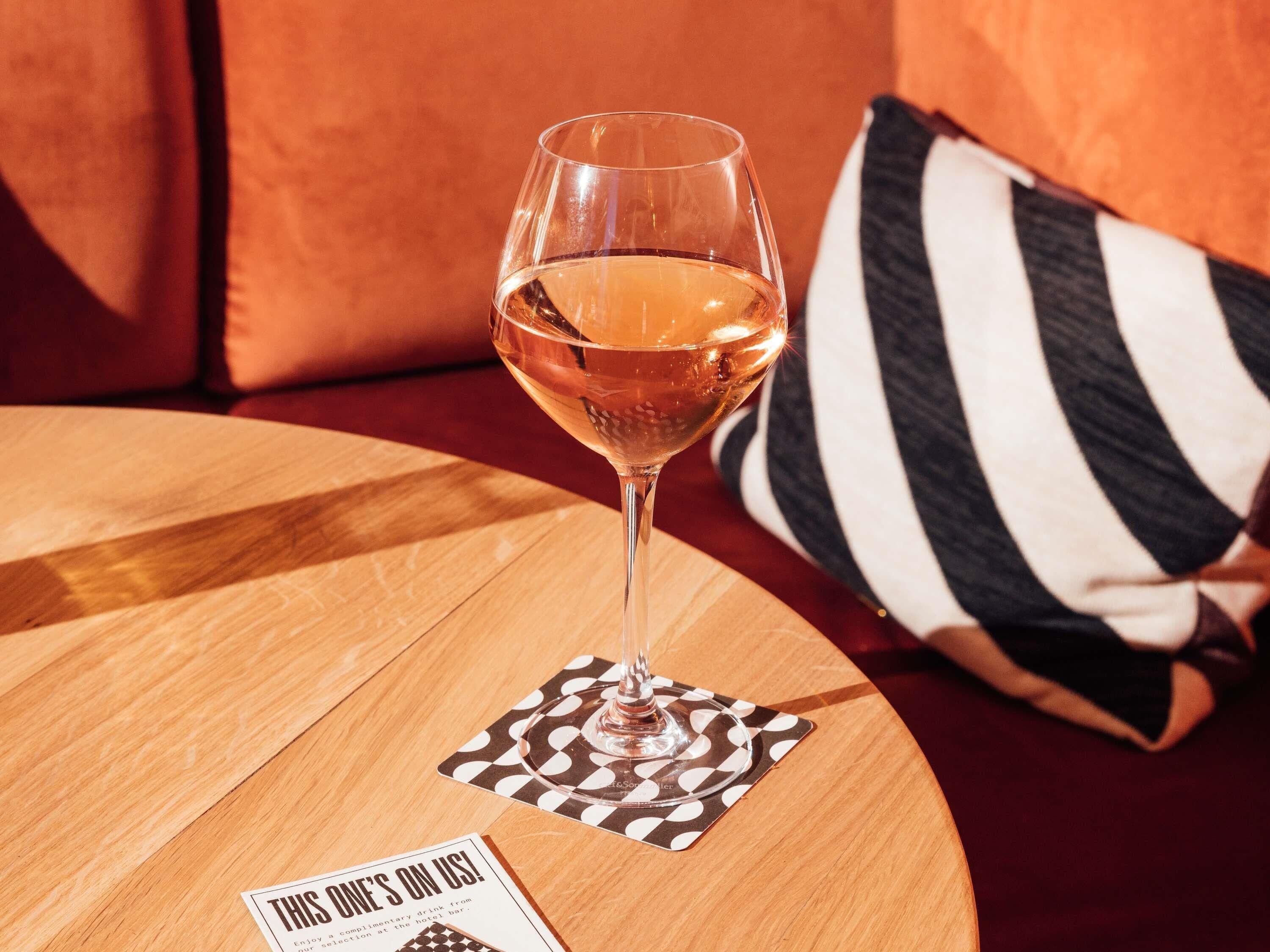 Un bicchiere di vino bianco su un tavolo rotondo di legno con un sottobicchiere a scacchi, un cuscino a righe bianche e nere e un cartellino dei menu.