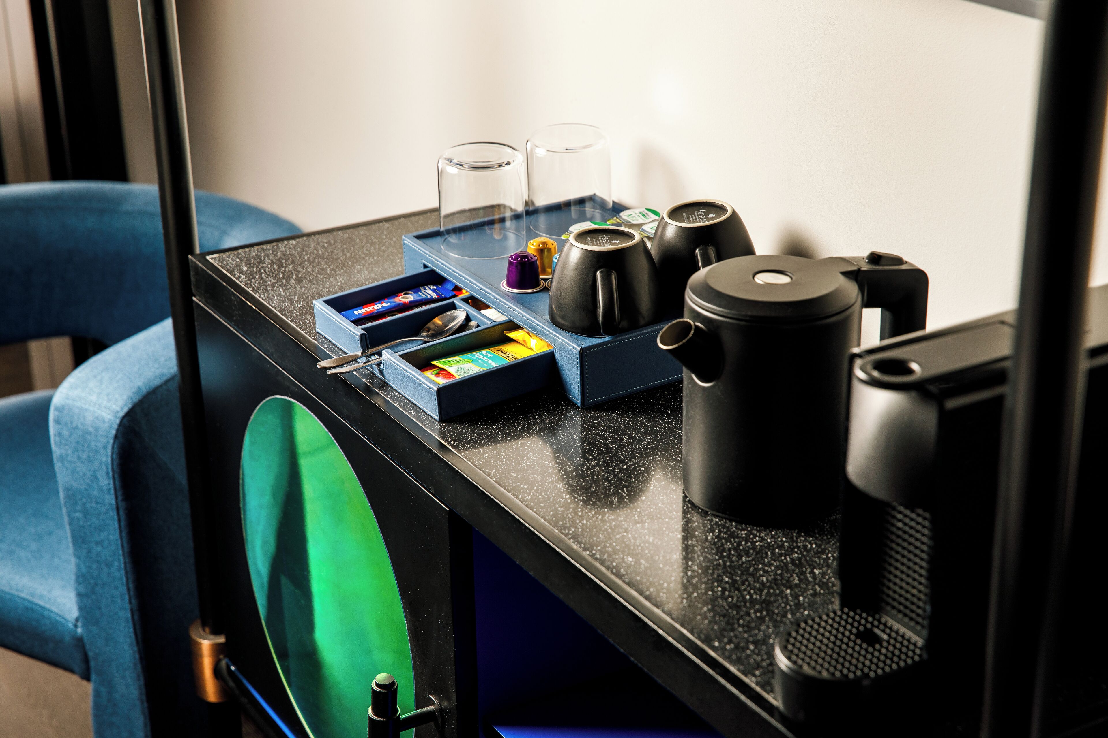 A coffee station with a coffee maker, cups, and a tray with coffee pods and a spoon.