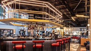 A modern bar area with a curved counter, red bar stools, and a variety of bottles and glasses. - TRIBE Manchester Airport (Manchester)