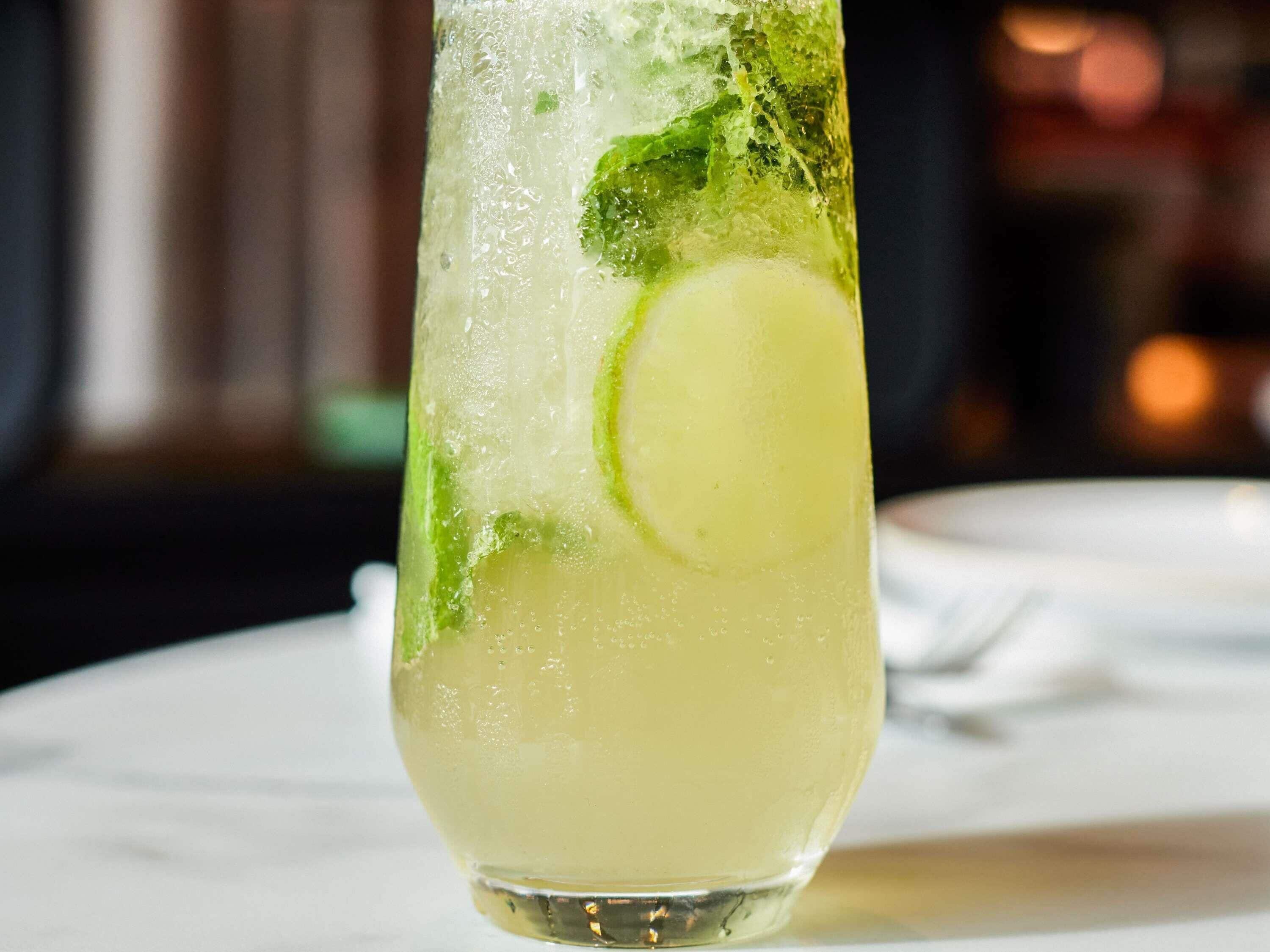 A tall glass with a lime slice and mint leaves, filled with a clear liquid, possibly a cocktail, on a white surface.