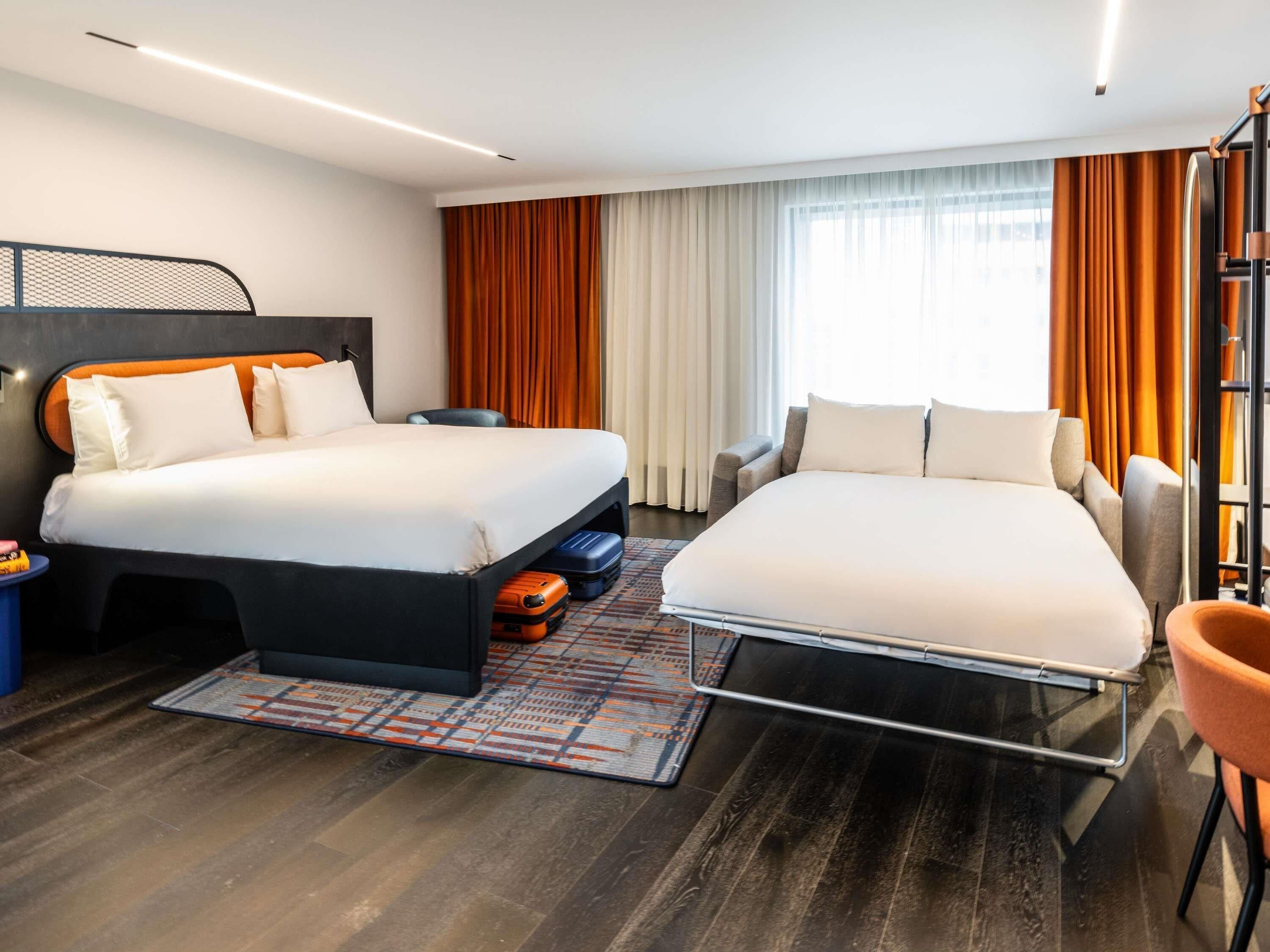 Una camera d'albergo con due letti, una scrivania, una sedia e un'ampia finestra con tende.