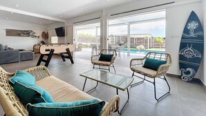 A spacious living area with a pool, wicker furniture, and a surfboard on the wall. - Lodge Wanalao - 4 rooms (Sainte-Anne)