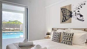 A bedroom with a bed, pillows, a nightstand, a lamp, and a view of a swimming pool. - Lodge Wanalao - 4 rooms (Sainte-Anne)
