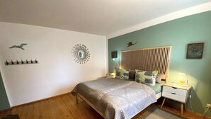 Un dormitorio con cama, piso de madera, ganchos en la pared, un espejo decorativo, una mesita de noche con lámpara y una alfombra pequeña.
