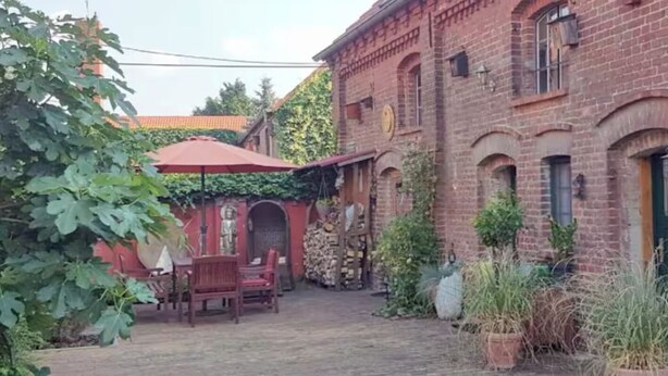 Un patio con un edificio de ladrillo, una banca de madera, un paraguas rojo y plantas en macetas.
