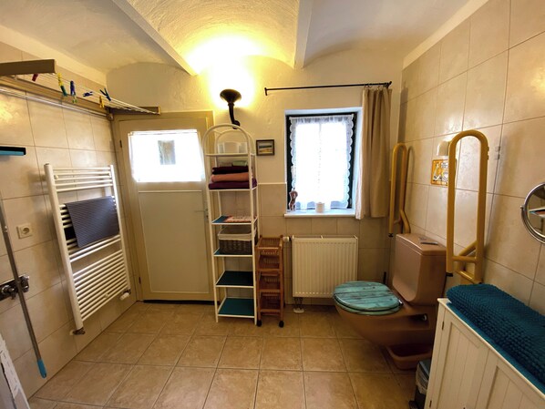 A compact bathroom with a white radiator, a window with lace curtains, a towel rack, and a shelf with folded towels. - Strodehne apartment, barrier-free, child-friendly (Havelaue)