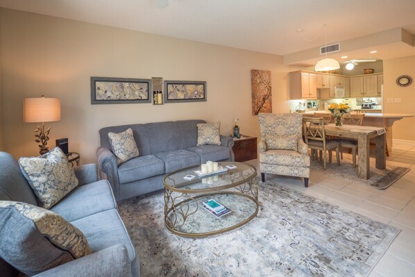 A living room with a grey sofa, patterned armchairs, a glass coffee table, and a dining area with a wooden table and chairs. - Beach Living at Regal Beach BLDE (George Town)