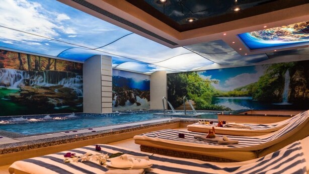 A spa area with a large indoor pool, reclining chairs, and a waterfall mural. - Royal Tulip Skikda (Filfila)